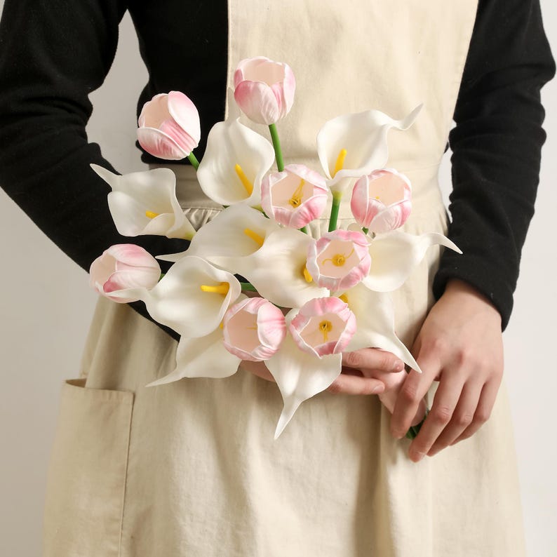 May include: A bouquet of artificial flowers featuring white calla lilies and pink tulips. The calla lilies have yellow centers, and the tulips are a soft pink. The flowers are held by a person wearing a black long-sleeve shirt and a beige apron.