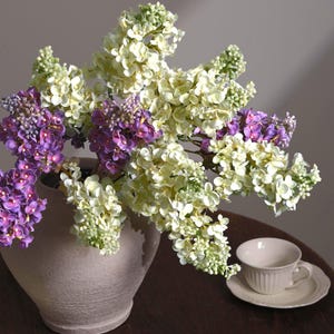 May include: A bouquet of artificial lilac flowers in shades of cream, white, and purple arranged in a textured, light beige ceramic vase. Beside the vase sits a matching cream-colored teacup and saucer with a ribbed, vertical pattern.
