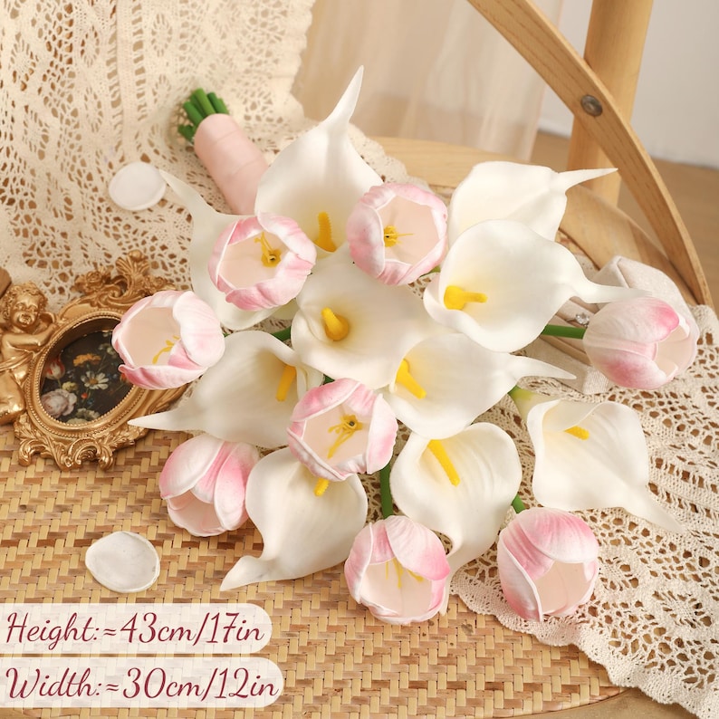May include: A bouquet of white calla lilies and pink tulips. The flowers are arranged on a woven surface. The bouquet is 17 inches high and 12 inches wide.
