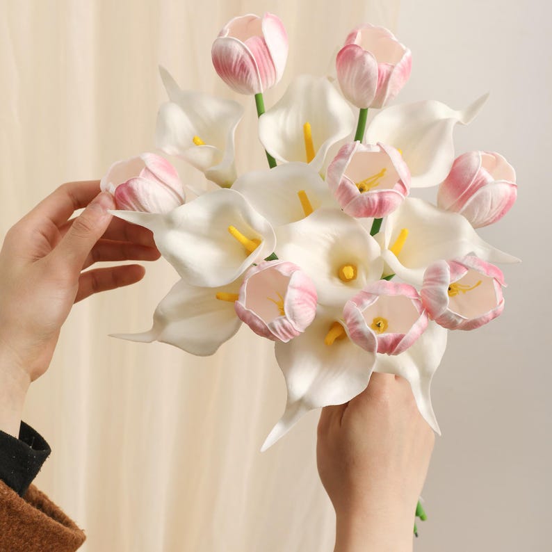 May include: A bouquet of white calla lilies and pink tulips. The calla lilies have yellow centers, and the tulips are a soft pink. The flowers are held in a person's hands, creating a visually appealing arrangement.