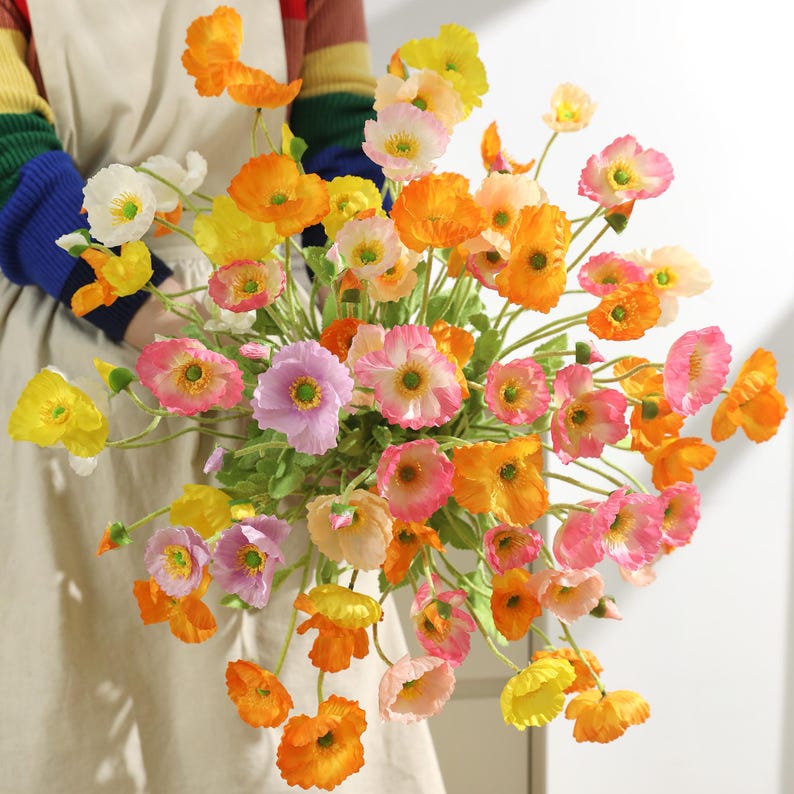 May include: A colourful bouquet of artificial poppies in shades of yellow, orange, pink, and white. The flowers have green stems. The poppy blooms are in various stages of opening, held against a neutral background.
