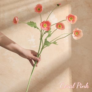 May include: A coral pink artificial flower stem with multiple blooms and green leaves. The flowers have a yellow centre and a delicate, layered petal design. The stem is held by a hand, and the text "Coral Pink" is visible.