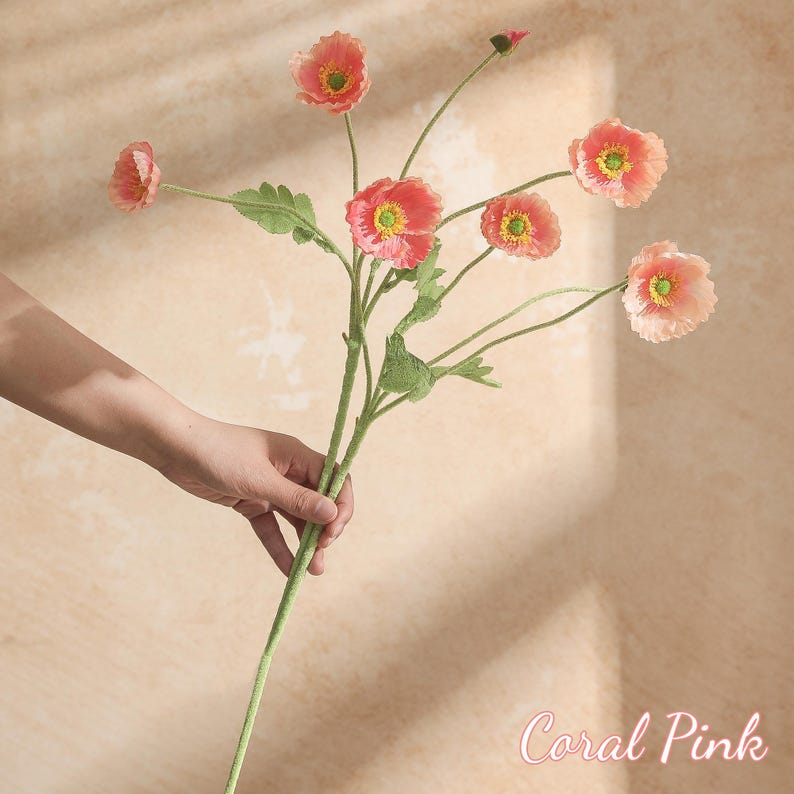May include: A coral pink artificial flower stem with multiple blooms and green leaves. The flowers have a yellow centre and a delicate, layered petal design. The stem is held by a hand, and the text "Coral Pink" is visible.