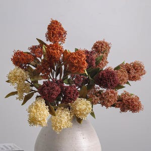 Könnte beinhalten: Ein Strauß künstlicher Blumen in einer hellen Vase, bestehend aus Büscheln kleiner, strukturierter Blüten in Creme, gebranntem Orange, tiefem Burgunderrot und Rosttönen, durchsetzt mit dunkelgrünen Blättern an dünnen Stielen.