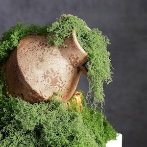 May include: A textured, light brown ceramic pitcher with a weathered, mottled finish, partially covered and overflowing with vibrant green preserved moss that cascades down the sides and pools around the base of the vessel.