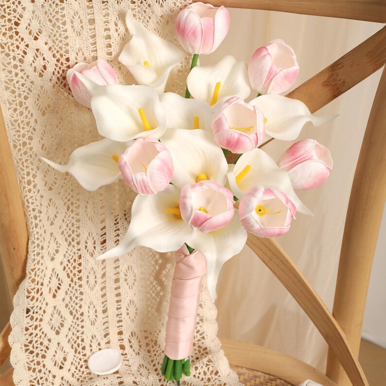 May include: A bouquet of white calla lilies and pink tulips tied with a pink ribbon. The flowers are arranged in a natural, elegant style, perfect for a wedding or special occasion. The bouquet is set against a light-colored background.