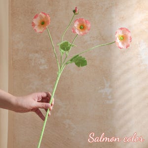 May include: An artificial flower stem in salmon pink with three open blooms and a bud. The flowers have a ruffled edge and a yellow centre. The stem is green with green leaves. The text "Salmon color" is in the bottom right corner.