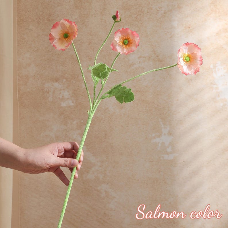 May include: An artificial flower stem in salmon pink with three open blooms and a bud. The flowers have a ruffled edge and a yellow centre. The stem is green with green leaves. The text "Salmon color" is in the bottom right corner.
