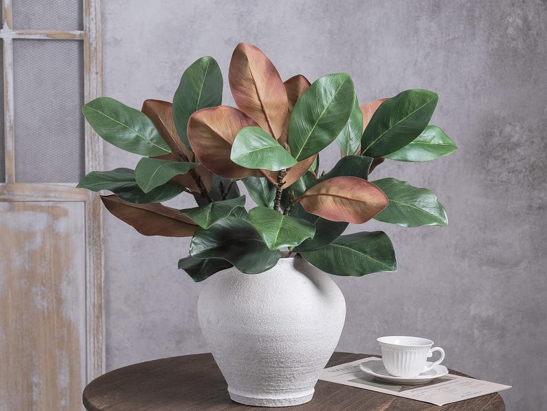 May include: A white ceramic vase with a textured surface containing a faux magnolia plant. The plant features large, glossy green leaves and several brown, copper-toned leaves. The vase is round with a narrow base and a wide, curved body.