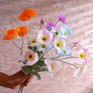 Könnte beinhalten: Ein Strauß künstlicher Mohnblumen in verschiedenen Farben, darunter Orange, Hellrosa, Weiß, Hellblau und Violett. Jede Blume hat eine markante gelbe Mitte und ist an einem dünnen grünen Stiel mit grünen Blättern befestigt.