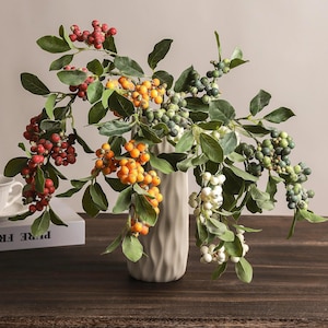 May include: A tall, beige, vertically ribbed ceramic vase holds several artificial branches featuring clusters of berries in shades of red, orange, white, and muted green, interspersed with realistic green leaves on thin, flexible brown stems.