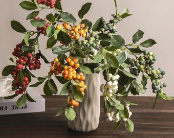 May include: A tall, beige, vertically ribbed ceramic vase holds several artificial branches featuring clusters of berries in shades of red, orange, white, and muted green, interspersed with realistic green leaves on thin, flexible brown stems.