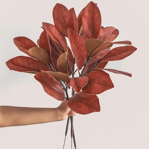 May include: A bouquet of artificial magnolia branches featuring deep rust-coloured, oval-shaped leaves with a smooth texture. The stems are dark brown and slender, branching out to support the dense arrangement of foliage in a fan-like display.
