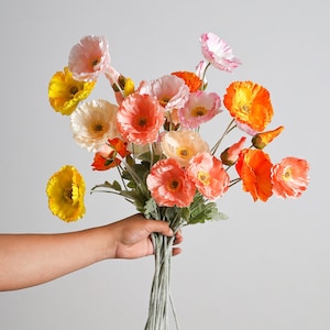 May include: A bouquet of artificial poppy flowers in shades of yellow, orange, peach, and light pink. The flowers feature delicate, ruffled petals and yellow centres, attached to thin green stems with small green leaves throughout the arrangement.