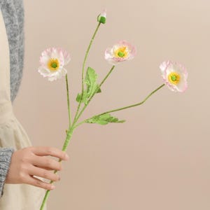 May include: A stem of artificial poppy flowers with three open blooms and a bud. The flowers have white petals with pink edges and yellow centres. The stem is green with green leaves.