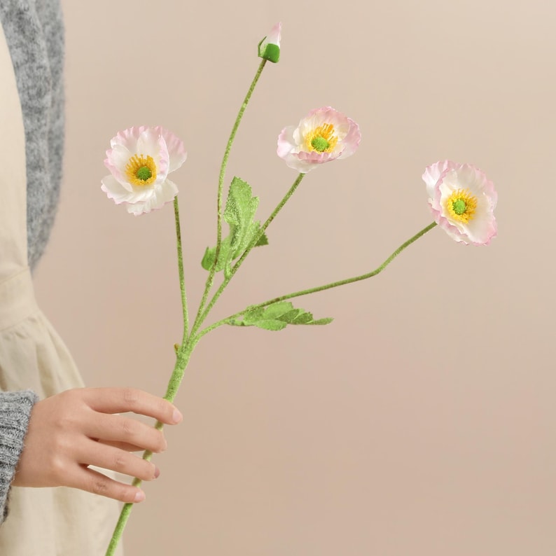 May include: A stem of artificial poppy flowers with three open blooms and a bud. The flowers have white petals with pink edges and yellow centres. The stem is green with green leaves.