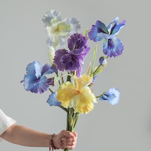 May include: A bouquet of artificial iris flowers featuring a variety of colors including deep purple, bright yellow, soft blue, and pale white. The flowers have ruffled petals and green stems, arranged together in a cluster with several buds.
