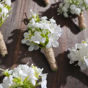 Könnte beinhalten: Mehrere weiße Blumen-Boutonnieres mit grünen Akzenten sind auf einer Holzoberfläche angeordnet. Jede Boutonniere besteht aus weißen Blüten und ist mit Bindfaden umwickelt. Die Anordnung deutet auf ein Hochzeits- oder besonderes Ereignis hin.