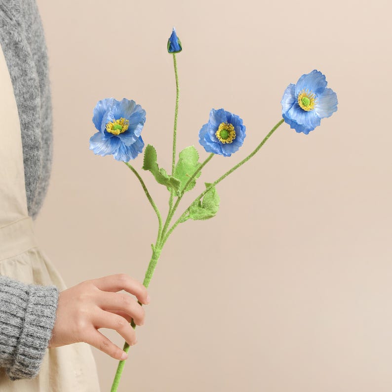May include: A stem of artificial blue poppy flowers with yellow centres and green leaves. The flowers are in full bloom, with one bud and a few green leaves. The stem is held by a person.