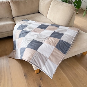 Könnte beinhalten: Eine Patchwork-Decke mit einem geometrischen Muster aus Quadraten und Rechtecken in Grau-, Beige- und Weißtönen. Die Decke liegt auf einem beigen Sofa und einer Ottomane und schafft eine gemütliche und einladende Atmosphäre. Maße: ca. 150 cm x 200 cm.