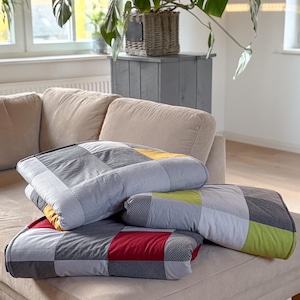 Könnte beinhalten: Ein Stapel von vier quadratischen Patchwork-Kissen in Grau-, Gelb-, Rot- und Grüntönen, die auf einem beigen Sofa liegen. Die Kissen haben verschiedene Muster, darunter Karos und Streifen, und sind in einem lässigen Haufen angeordnet.