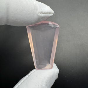 以下が含まれることがあります： 台形型の、薄いピンク色のファセットカットの宝石。手袋をはめた手が暗い背景に宝石を持っています。石のファセットは光を捉え、その透明度と色を示しています。
