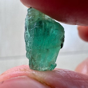 Puede incluir: Una esmeralda en bruto sin pulir con un tono verde vibrante y translúcido. El cristal muestra una estructura columnar hexagonal natural con estrías verticales visibles, bordes irregulares e inclusiones internas en toda su superficie.