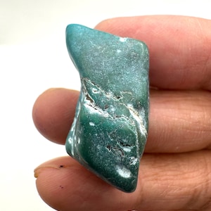 Puede incluir: Una pequeña piedra pulida de forma irregular con un color verde azulado profundo y verde bosque. La superficie presenta una textura lisa y pulida con fisuras naturales visibles, vetas minerales blancas y pequeñas hendiduras.