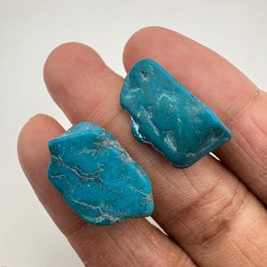 May include: Two irregular, tumbled turquoise stones with a vibrant blue-green colour and natural white veining. The stones have a smooth, polished texture and display unique, organic shapes with varied surface contours and edges.