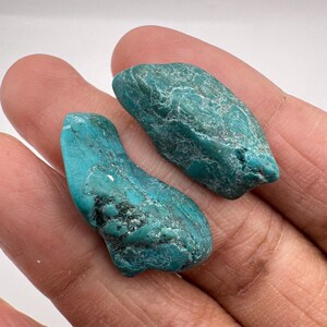 May include: Two raw, unpolished turquoise stones with irregular, organic shapes. The stones feature a vibrant teal-blue colour with natural dark veining and a matte, textured surface. Each piece displays unique geological formations and variations.