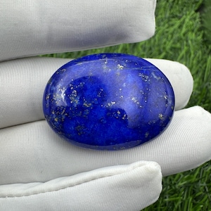 Puede incluir: Una piedra preciosa de lapislázuli de forma ovalada con una superficie pulida y lisa. La piedra presenta un color azul profundo y vibrante con inclusiones de pirita de color oro metálico dispersas por toda la superficie.