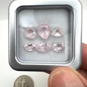Può includere: Sei pietre preziose sfaccettate rosa pallido disposte in due file all'interno di una custodia quadrata color argento con coperchio trasparente. Le pietre presentano vari tagli, tra cui forme a pera, triangolo e ovale. È visibile una piccola moneta d'argento.