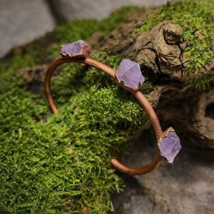 May include: A copper-colored bracelet featuring three raw, light purple amethyst crystals. The bracelet is set against a backdrop of green moss and a piece of driftwood, highlighting the natural elements of the jewelry.