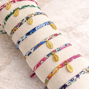 May include: A collection of seven fabric bracelets featuring various floral patterns in colors including pink, green, blue, and yellow. Each bracelet is adorned with a small, oval-shaped gold-toned metal charm depicting a religious figure.