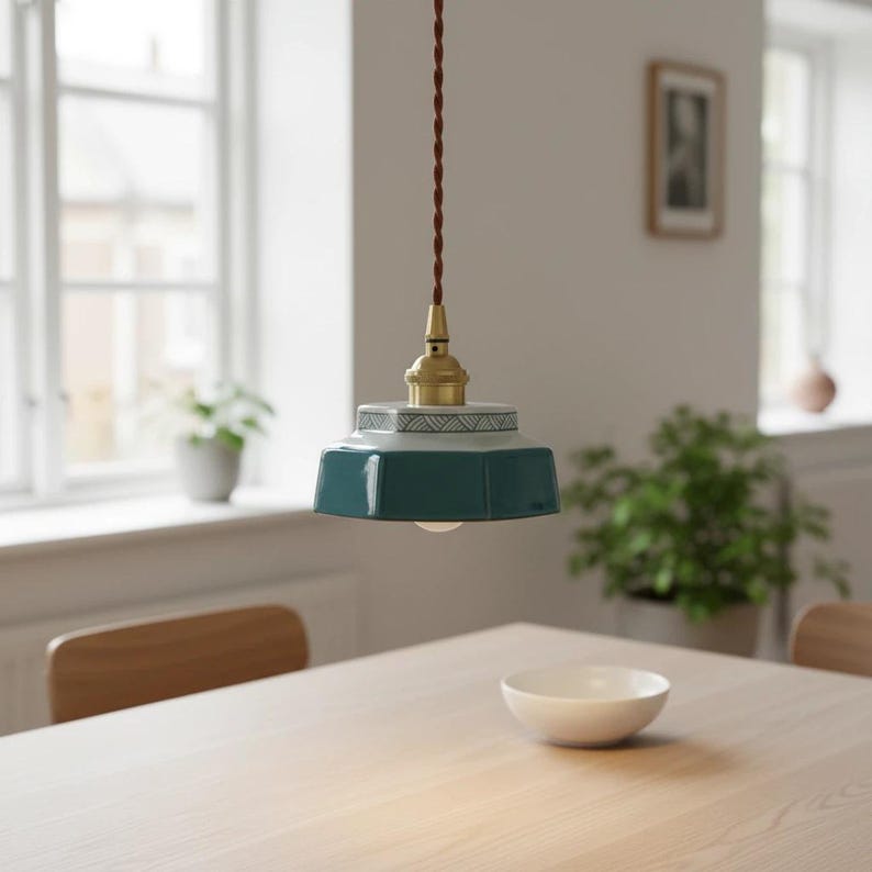 May include: A teal and white pendant light fixture with a geometric design. The light hangs from a brown cord and has a brass-colored base. The light is hanging above a wooden table, with a white bowl on the table.