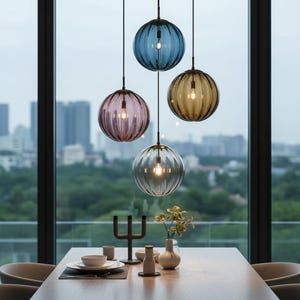 May include: Four spherical pendant lights with ribbed glass shades in blue, pink, amber, and grey hang above a dining table. Each light has a black cord and a warm bulb. The lights are suspended in front of a large window overlooking a cityscape.