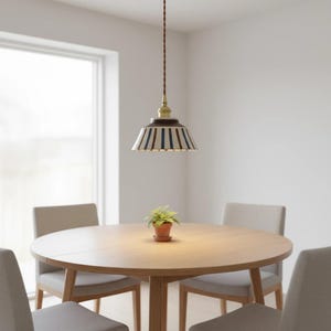 May include: A pendant light fixture with a striped lampshade in white, navy blue, and brown hangs above a round wooden table. The fixture has a brass-colored top and a brown cord. Four gray chairs surround the table, and a small potted plant sits on the table.