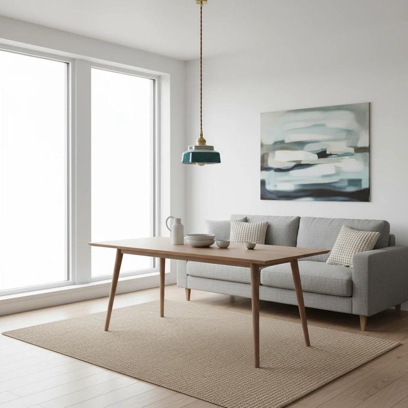 May include: A modern dining room scene featuring a light wood dining table with angled legs, a gray sofa, and a woven rug. A teal and gold pendant light hangs above the table. An abstract painting and large windows complete the room.
