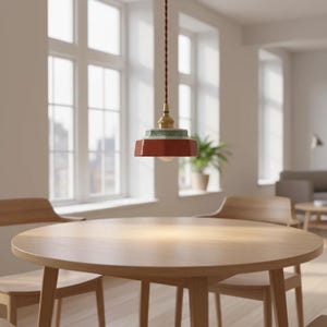 May include: A modern pendant light with a red, geometric shade and a brass fixture hangs above a round wooden table. The light has a green and white patterned accent. The room has large windows and wooden chairs.
