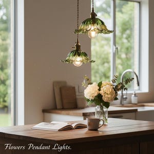 May include: Two green and gold flower-shaped pendant lights hanging from the ceiling. The lights are above a wooden table with a book, a mug, and a vase of white flowers. The text "Flowers Pendant Lights" is at the bottom.
