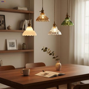 May include: Four pendant lights with scalloped glass shades in white, amber, clear, and green hang above a wooden dining table. The lights have brass-coloured fixtures and are suspended from the ceiling. A vase with greenery and a coffee cup are on the table.