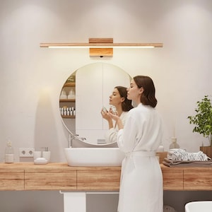 Puede incluir: Una lámpara de tocador de madera con una base rectangular de madera y una barra de luz horizontal larga. La luz está montada sobre un espejo redondo en un baño. El baño tiene un tocador de madera y un lavabo blanco.