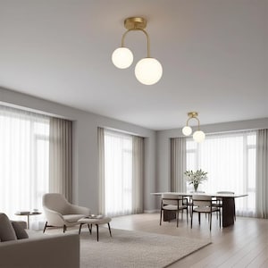 May include: A modern, gold-toned ceiling light fixture with two spherical, frosted glass globes. The fixture is suspended from a curved arm, creating a minimalist design. The light is installed in a living room with neutral tones and large windows.