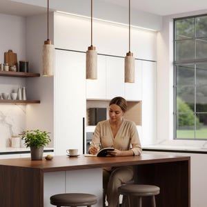 May include: A modern kitchen features three cylindrical pendant lights with beige stone-like shades and wooden accents. A person sits at a dark wood island, writing in a notebook. The kitchen has white cabinets, a large window, and a small potted plant.