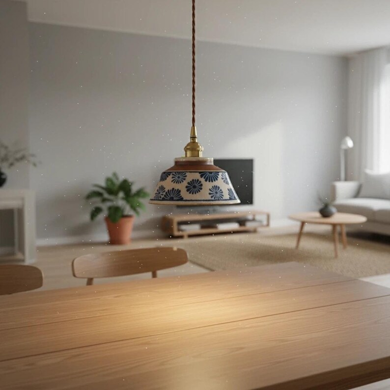 May include: A pendant light fixture with a floral patterned lampshade in blue and cream. The light has a brass fitting and a brown cord. The fixture hangs above a wooden table in a well-lit room, suggesting a dining or living area.