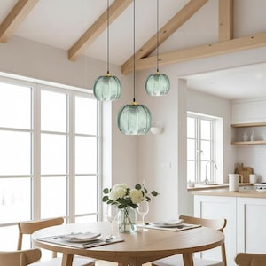 May include: Three pendant lights with teal glass shades hang above a round wooden dining table. The shades have a ribbed design and gold-colored accents. The room features large windows, wooden beams, and a vase of white flowers.