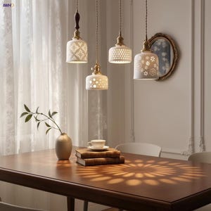 Peut inclure: Quatre lampes suspendues en céramique blanche avec des motifs découpés pendent au-dessus d'une table en bois. Les lampes ont des accents en laiton et sont suspendues par des cordons marron. Un vase avec une plante et une tasse de thé sont sur la table.