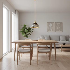 May include: A light wood dining table with four matching chairs, set in a bright room. A striped pendant light hangs above the table. A large green plant and a gray sofa complete the modern interior design.