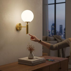 May include: A contemporary gold wall light with a round white glass shade. The light is mounted on a beige wall. A person's finger is touching a crystal switch. A small vase of dried flowers sits on a wooden surface.