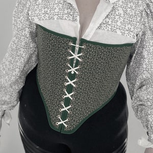 May include: A woman wearing a green and white corset with white lacing. The corset has a textured fabric and a pointed bottom. She is also wearing a white blouse with a black and white pattern and black trousers.